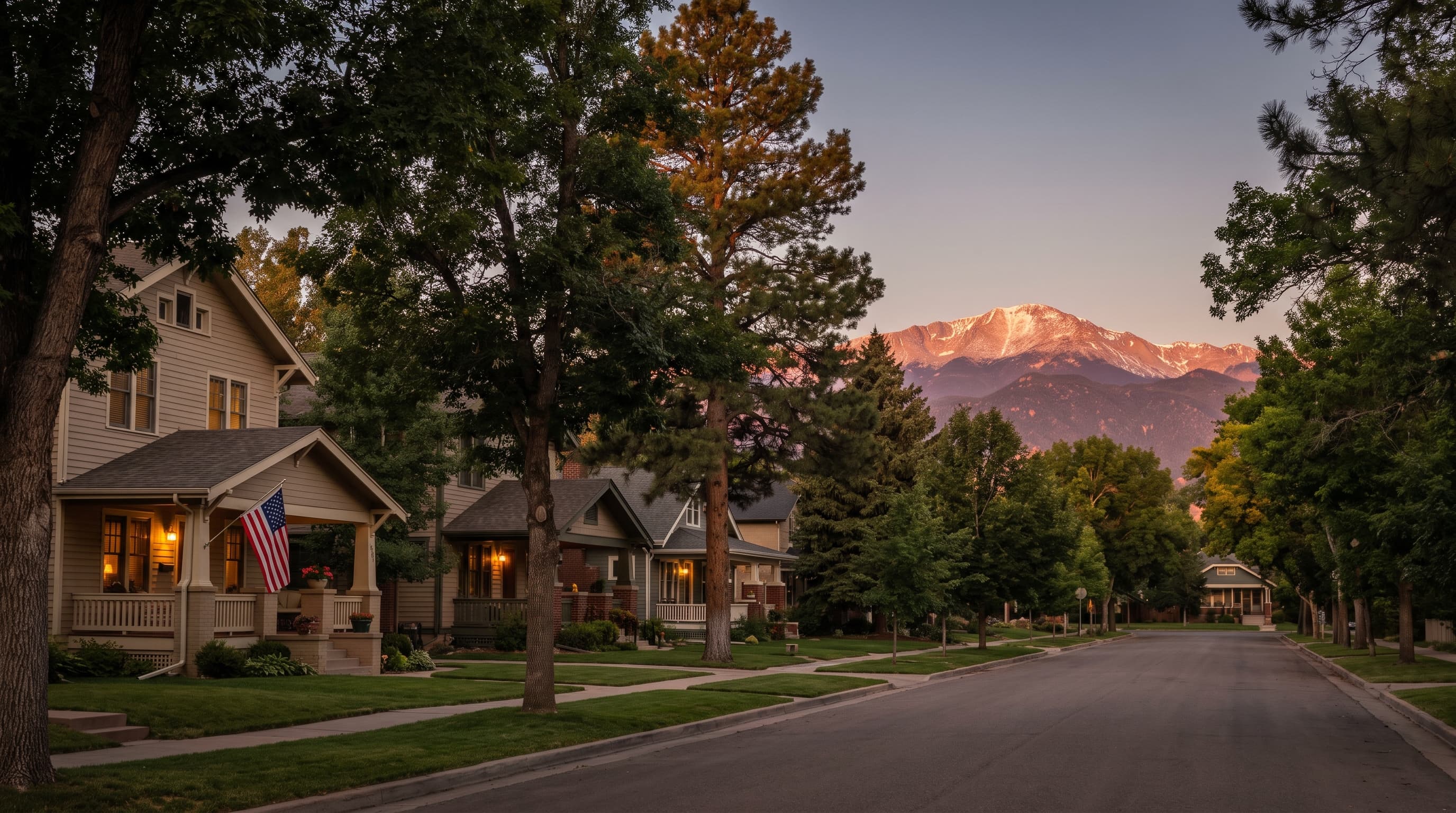 Tree-lined Colorado Springs neighborhood street at dusk with a flag on a porch and Pikes Peak in alpenglow